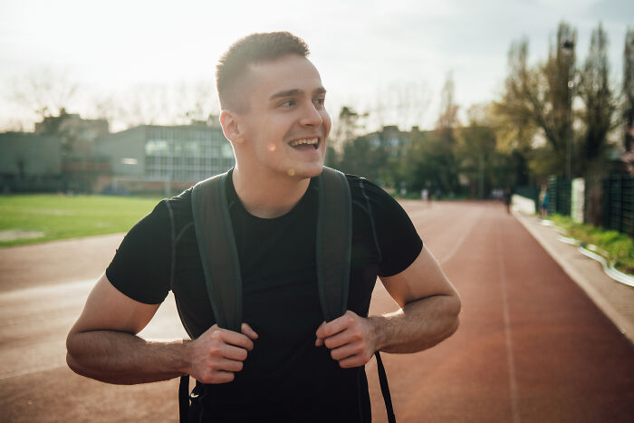 Young man wearing a black shirt and backpack smiling outdoors on a running track with clear sky beyond, sewer workers bizarre discoveries.