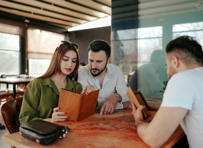 Three restaurant customers reading menus while sitting at a wooden table in a casual dining setting with servers nearby.