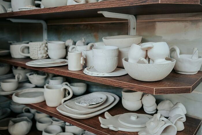 Shelves filled with unpainted ceramic pottery pieces drying in a workshop, showcasing handmade cups and plates.