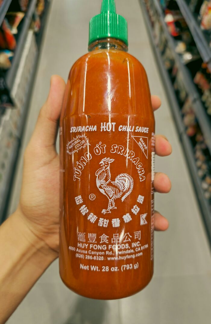 Hand holding a bottle of Sriracha hot chili sauce in a grocery store aisle, highlighting tattoo artist challenges.