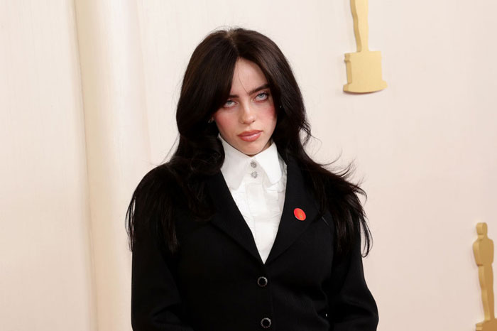 Billie Eilish wearing black suit and white shirt, looking serious amid fans outraged after Miami concert incident.