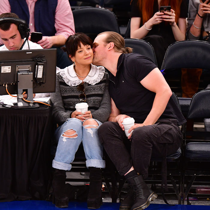 Lily Allen sitting courtside with a man whispering, both holding coffee cups, amid album and cheating controversy buzz.