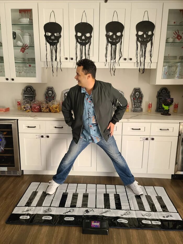 Man standing on oversized piano mat in Halloween-themed kitchen, showing creative transformation dedication to Tom Hanks faces.
