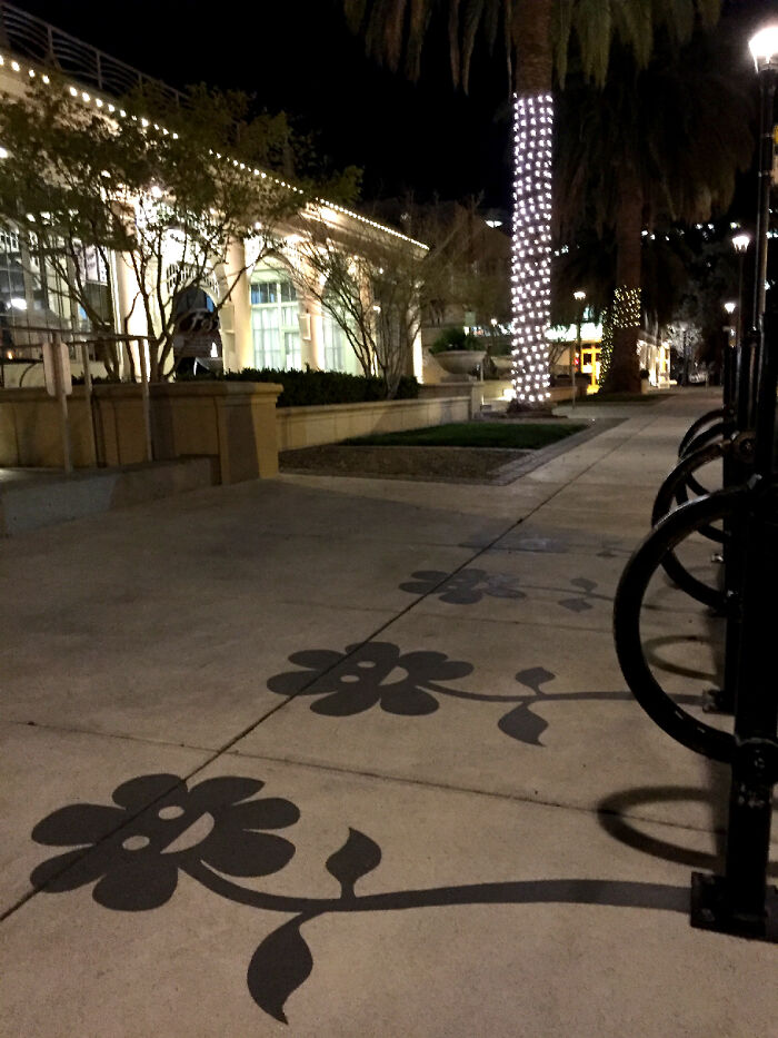 Black metal bike racks casting clever shadow art of smiling flowers on a concrete sidewalk in an urban setting.