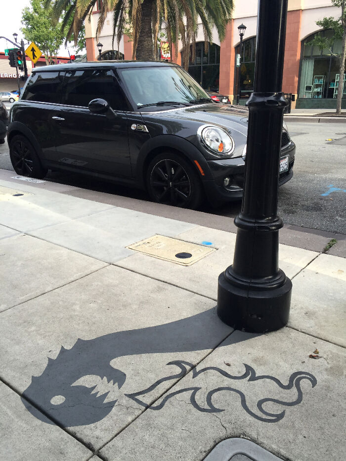 Street lamp shadow art installation on sidewalk creatively transforms shadow into a dragon breathing fire in clever shadow art.