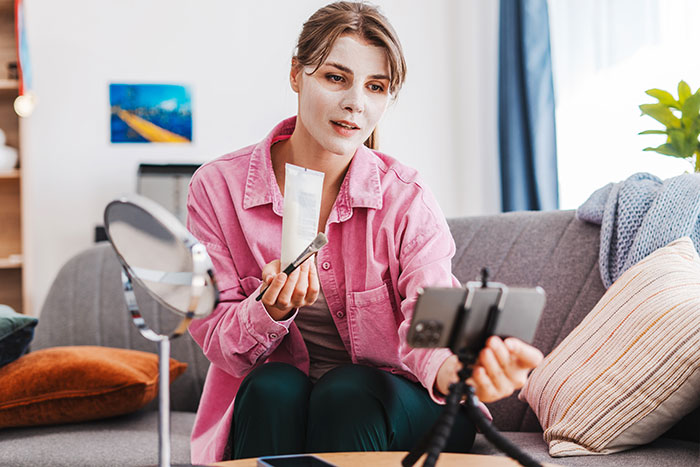 Gym influencer recording skincare video at home, showing product and makeup brush while filming on smartphone tripod.