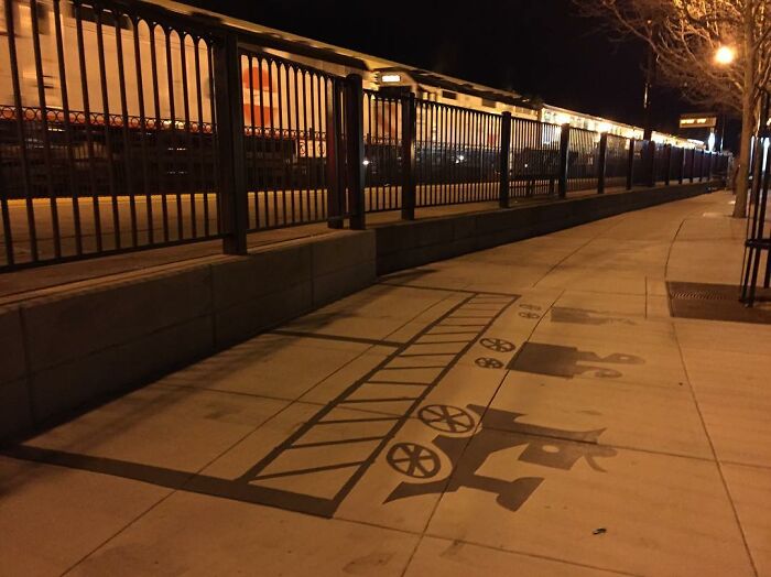 Sidewalk painted shadow art installation of a vintage train with a ladder, wheels, and silhouetted figures on a city street.