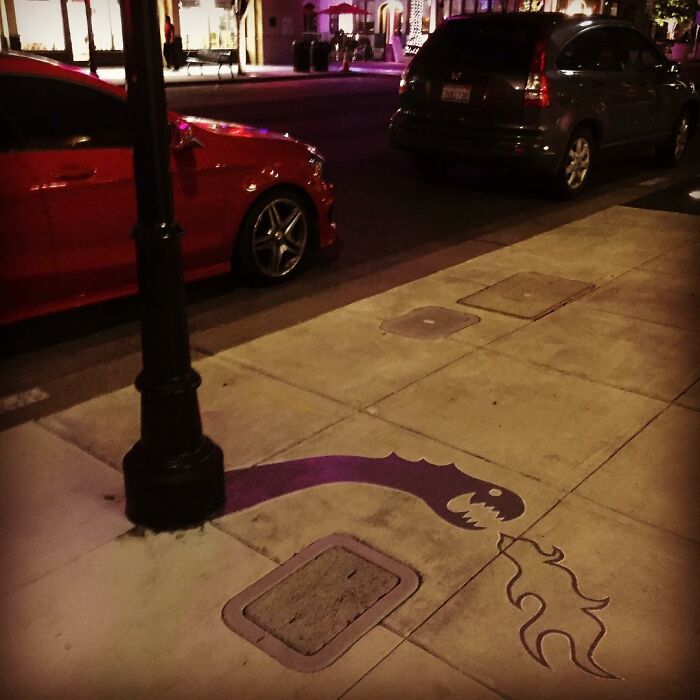 Street lamp shadow art installation on sidewalk creatively transforms shadow into a dragon breathing fire in clever shadow art.