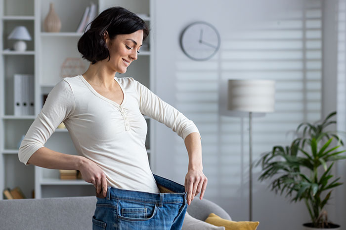 Woman smiling and holding oversized jeans, showing weight loss in a bright living room with clock and plants. Woman smiling and holding oversized jeans, showing weight loss in a bright living room with clock and plants.