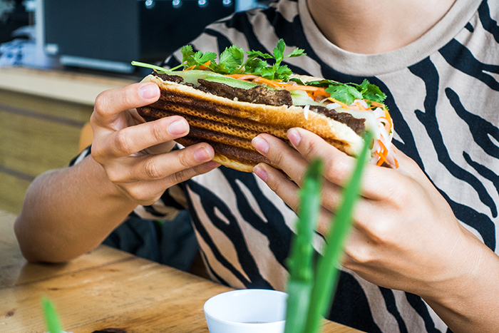 Person holding and about to eat a spicy chicken sandwich topped with fresh herbs and vegetables at a wooden table