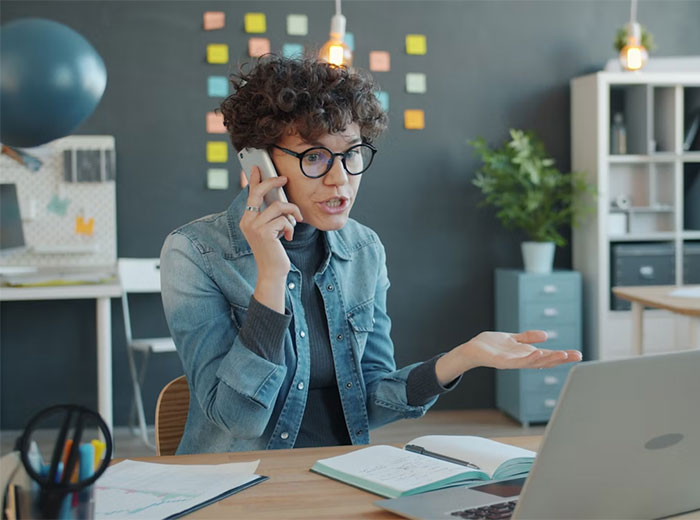 Woman with curly hair and glasses making a frustrated phone call about doctor refusing to rehire sister after costly mistake.