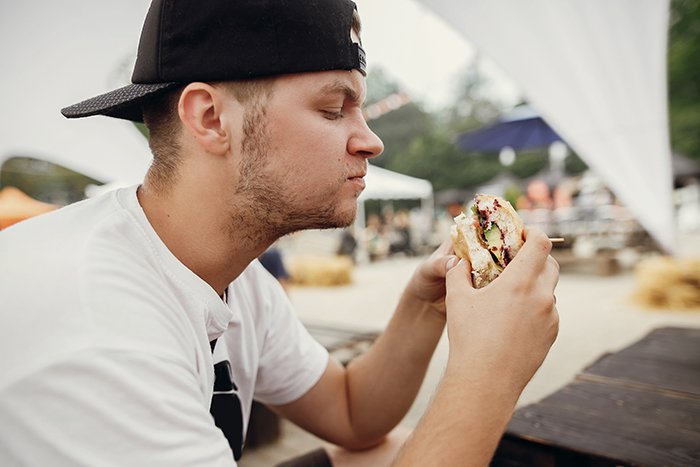 Young man wearing a cap tasting a spicy chicken sandwich outdoors at a casual picnic area on a cloudy day.
