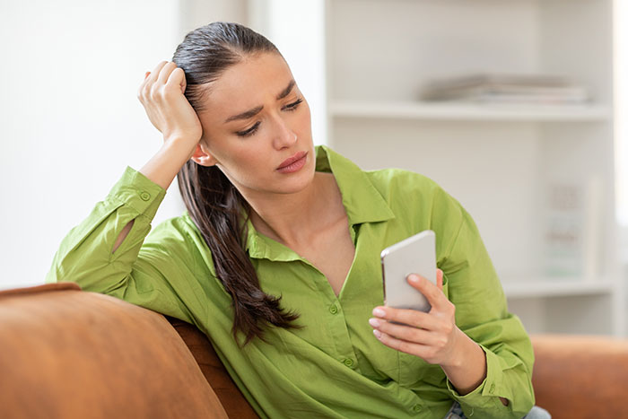 Woman looking surprised while holding phone on couch, relating to bumps into boyfriend&rsquo;s grown son wake-up call.