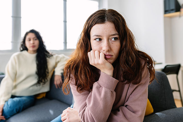 Young woman looking upset and distant on a couch while her sister sits behind her, reflecting a troubled family life. Young woman looking upset and distant on a couch while her sister sits behind her, reflecting a troubled family life.
