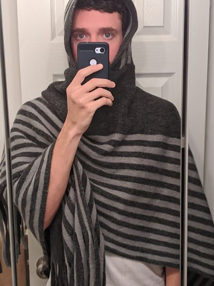 Man wearing a striped shawl taking a mirror selfie with a smartphone in a bathroom doorway.