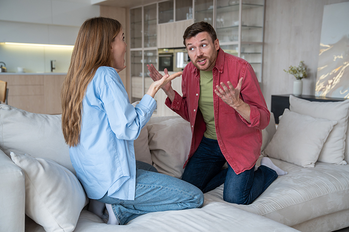 Woman and husband having a heated argument on couch about his Pok&eacute;mon card collection in a modern living room setting.