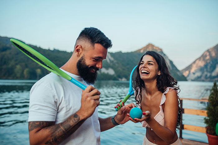 Couple playing with rackets and ball by the lake, woman staging apartment to impress a guy, sharing a joyful moment.