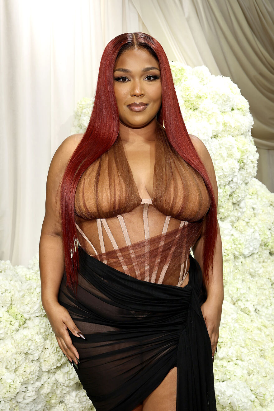 Lizzo posing in a sheer brown top and black skirt in front of white flowers, showcasing wellness evolution.