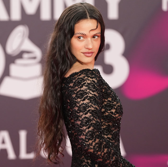 Pop star with dyed armpit hair posing at Paris Fashion Week, wearing a black lace dress and long wavy hair.