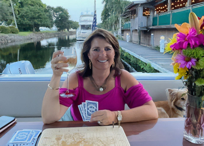 Woman writes her own obituary with humor, smiling while holding playing cards and a glass on a boat deck.