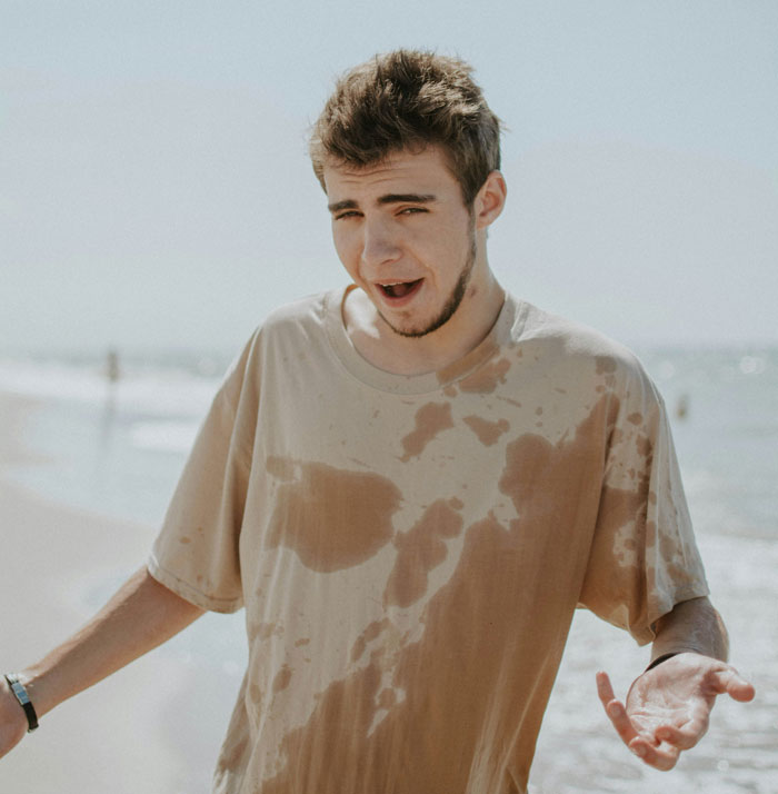 Young man with a wet shirt and confused expression at the beach, showing hilarious instances of common knowledge unawareness