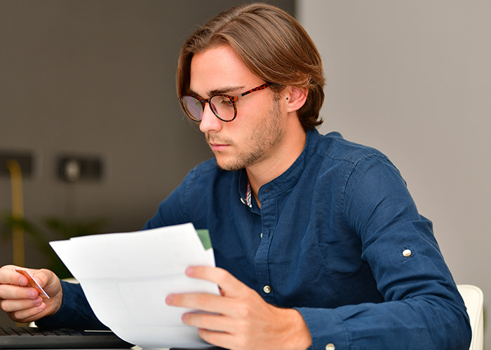Young accountant wearing glasses and a blue shirt, focused on reviewing financial documents at his desk.