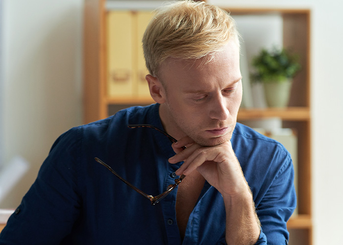 Young male accountant in blue shirt holding glasses, deep in thought while working in a modern office setting.