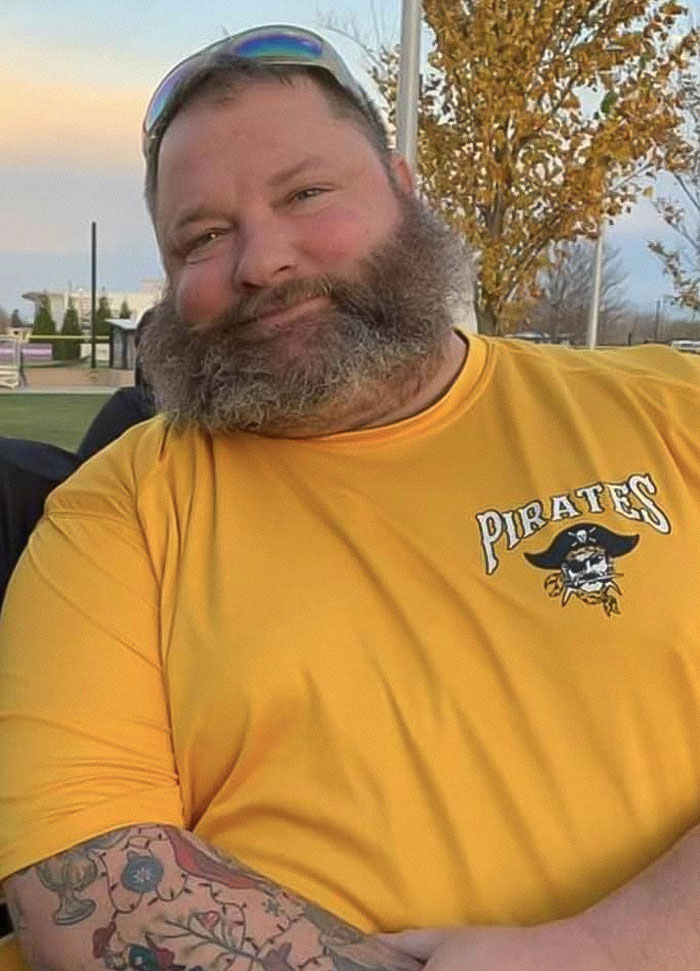 Morbidly obese man with a beard wearing a yellow Pirates shirt, outdoors with trees and a park in the background.