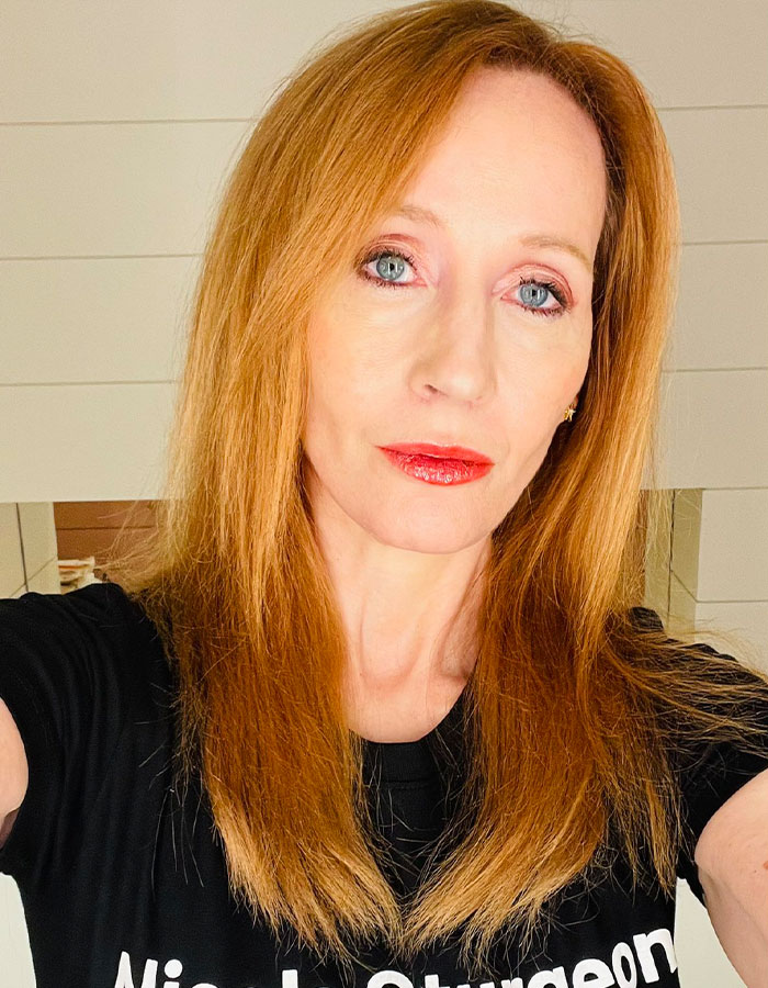 J.K. Rowling with red hair and red lipstick wearing a black shirt, posing indoors with a neutral expression.