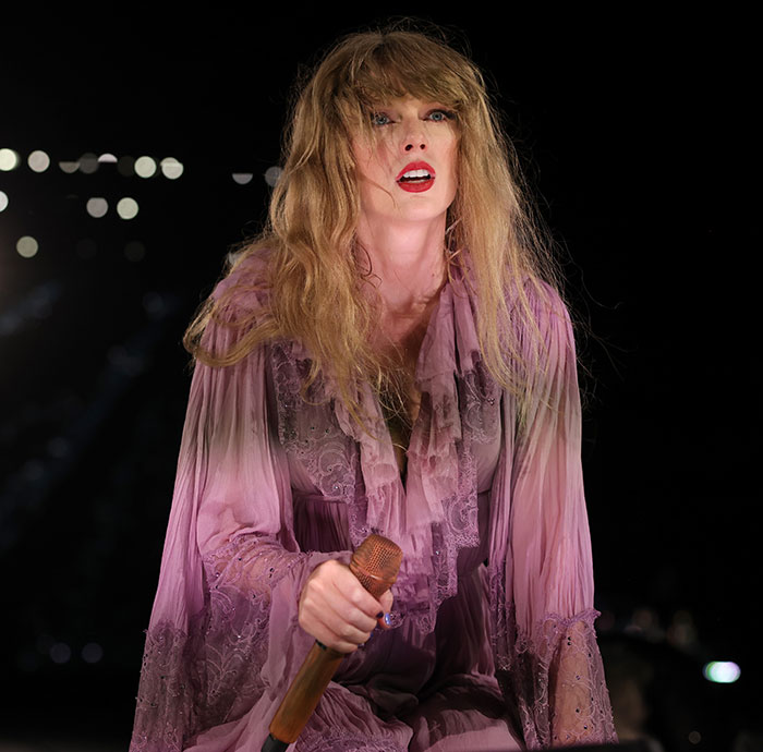 Taylor Swift performing on stage wearing a pink lace dress, holding a microphone under bright concert lights.