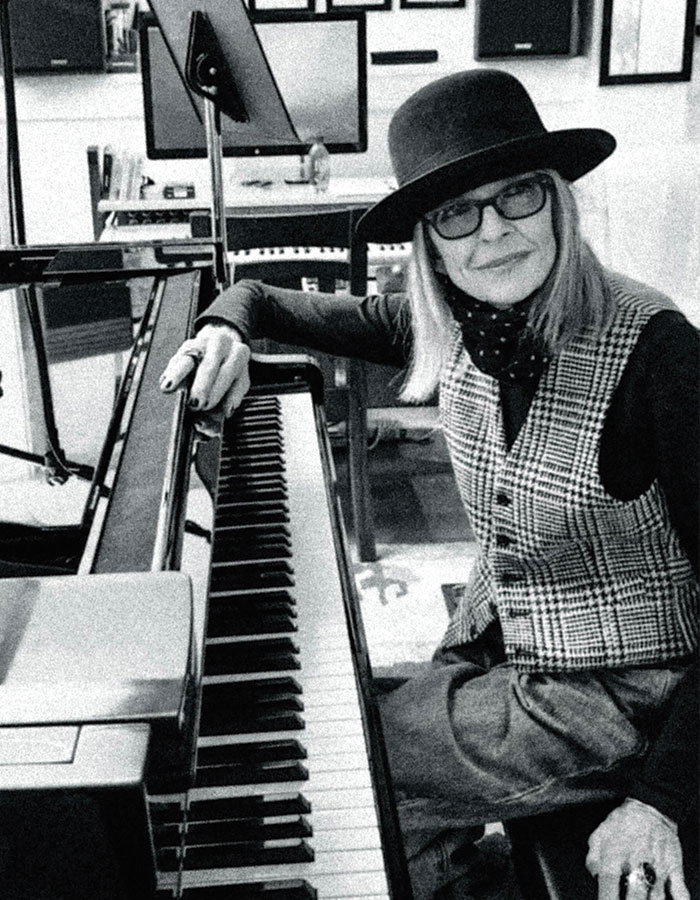 Diane Keaton seated at a piano, wearing a hat and glasses, in a room filled with musical equipment and art.