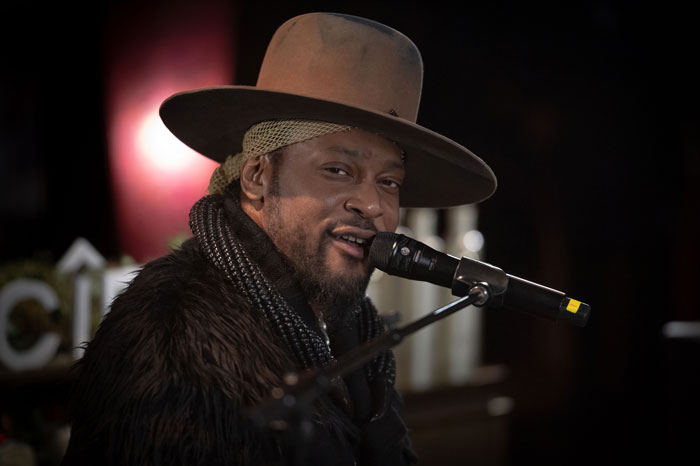 R&B legend D'Angelo singing into a microphone wearing a wide-brimmed hat and fur coat during a live performance.