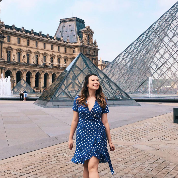 Pregnant Disney superfan smiling in a blue polka dot dress near glass pyramid structures at a historic museum courtyard.