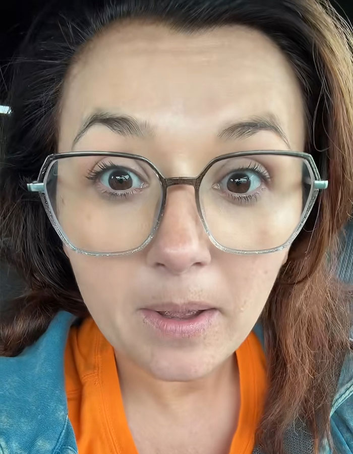 Woman with glasses and orange shirt showing shocked expression related to husband infidelity confrontation mid-flight.