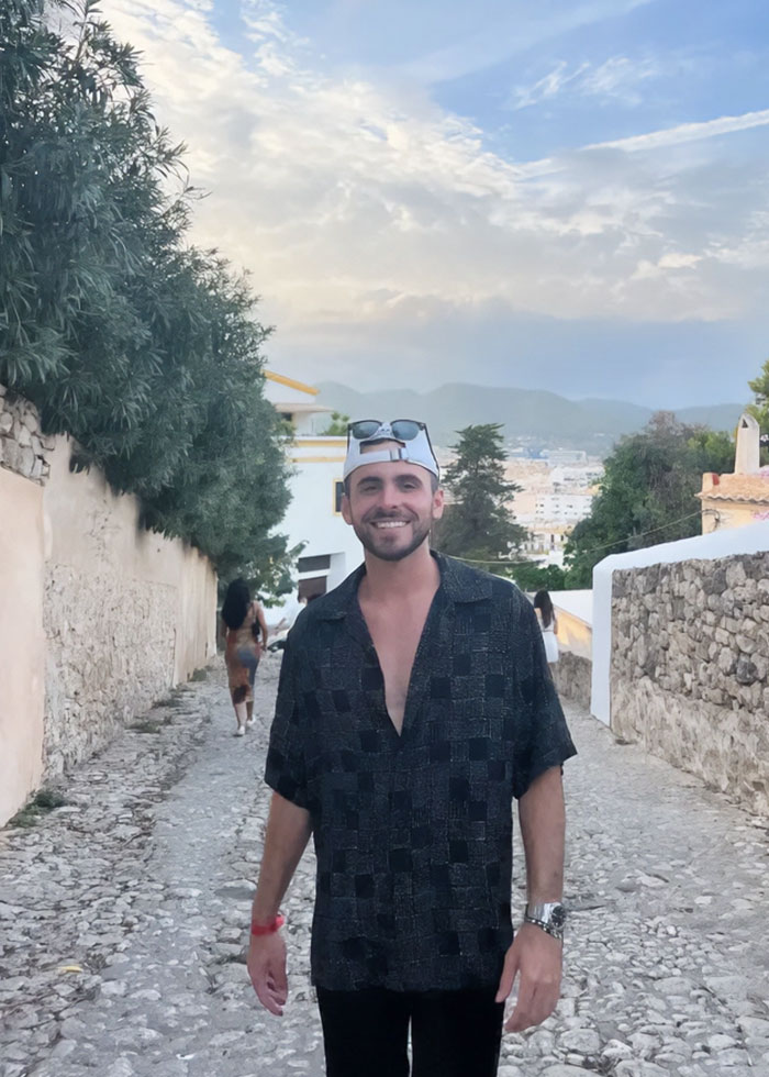 Young man smiling outdoors during hair transplant recovery, wearing a patterned shirt and white cap on a cobblestone street.