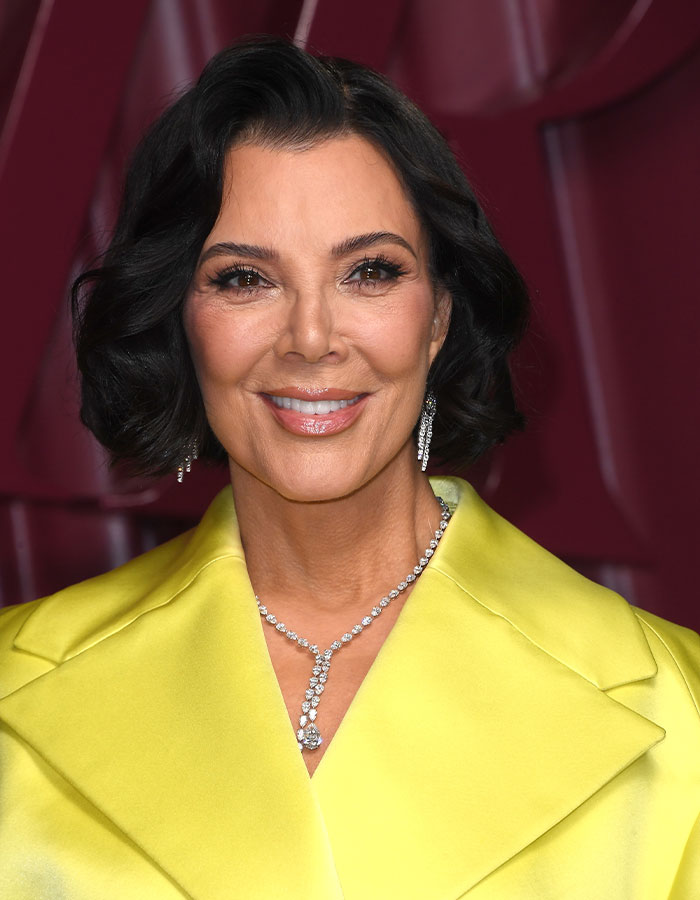 Kris Jenner smiling in a yellow outfit with diamond earrings and necklace at a public event with fans' wild reactions.
