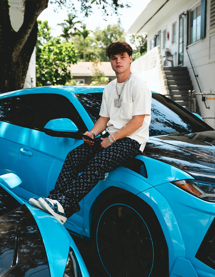Young man sitting on a blue car outdoors, reflecting style and attitude, related to adult star wedding vows and YouTube prankster split.