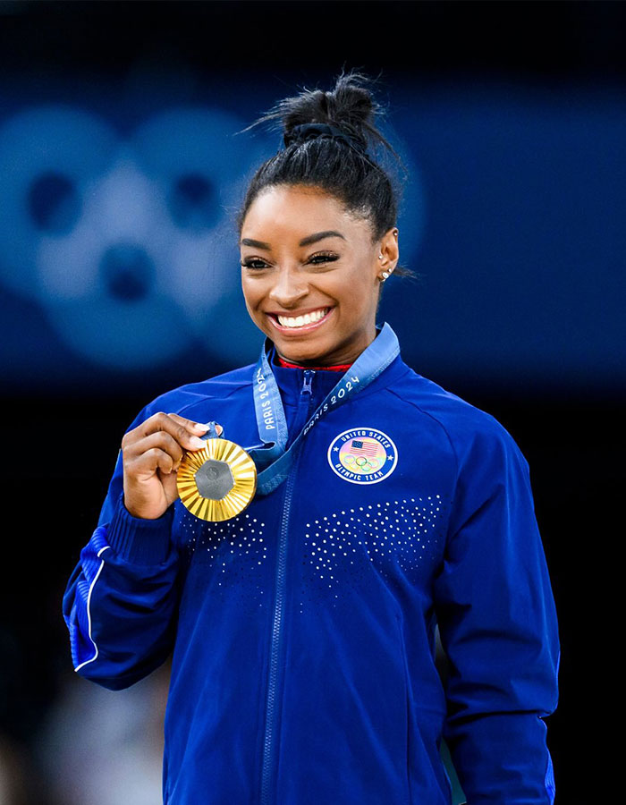 Simone Biles smiling and holding a gold medal at the Paris 2024 Olympics, representing the United States Olympic team.