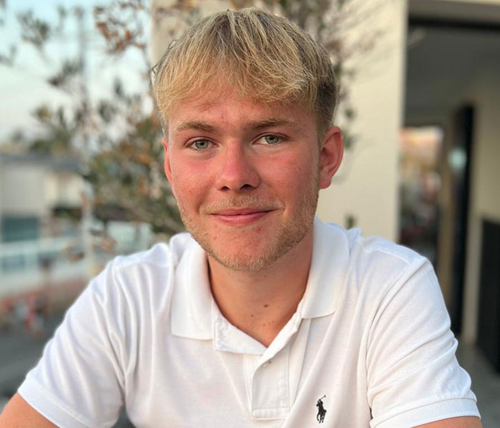 Young man with blonde hair wearing a white polo shirt, smiling outdoors after deciding to amputate his leg post rock concert.