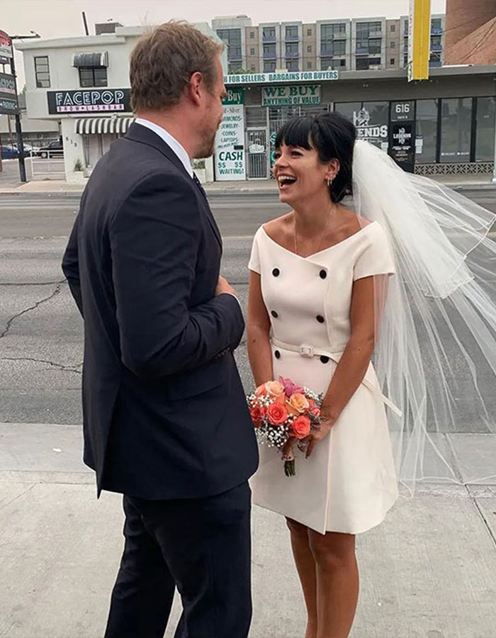 Lily Allen in a white dress and veil holding flowers, smiling and talking with a man in a dark suit outdoors.