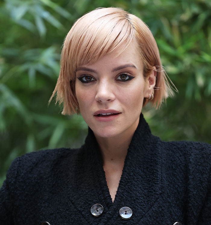 Close-up of a woman with short blonde hair wearing a black textured coat outdoors with greenery in the background, David Harbour friend.