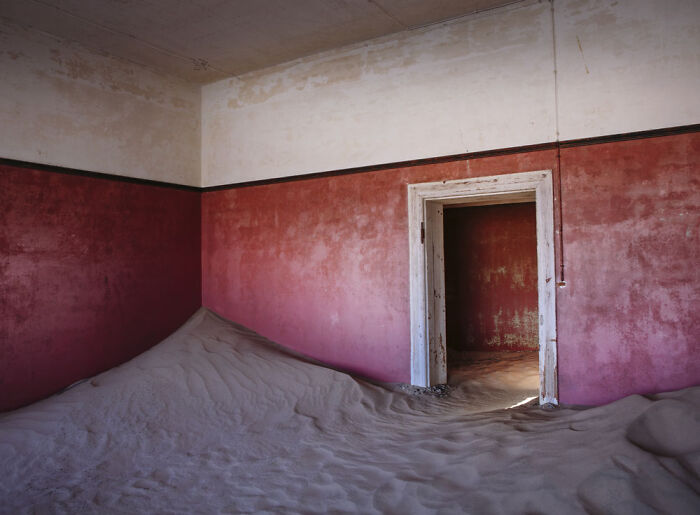 Desert sand filling an abandoned home with red walls, illustrating surreal sand castles engulfing deserted interiors.