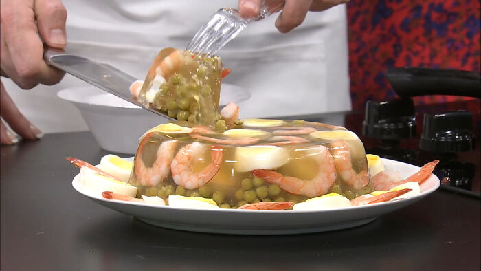 A slice of shrimp jello being served from a gelatin mold with peas, shrimp, and boiled eggs on a plate.