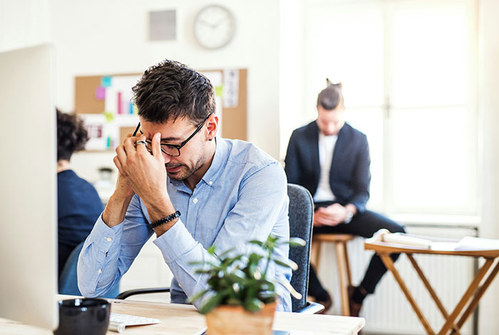 Frustrated employee at desk holding glasses, stressed after not checking email on public transit before work.