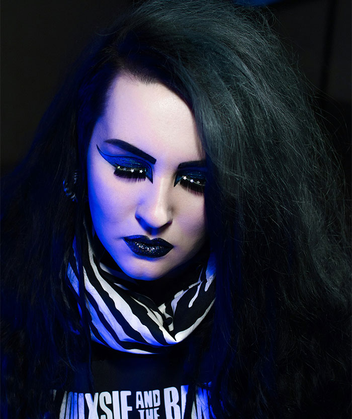 Woman with dramatic dark makeup and black hair looking down, reflecting on past bullying and emotional struggles.