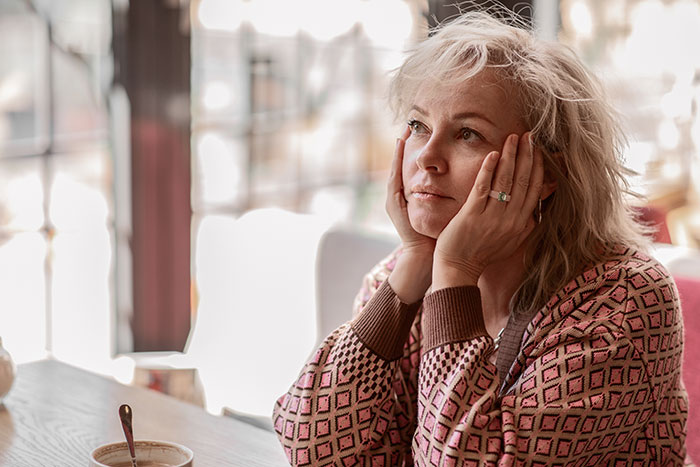 Woman sitting thoughtful by a window, wearing patterned sweater, reflecting complex emotions in a quiet indoor setting.