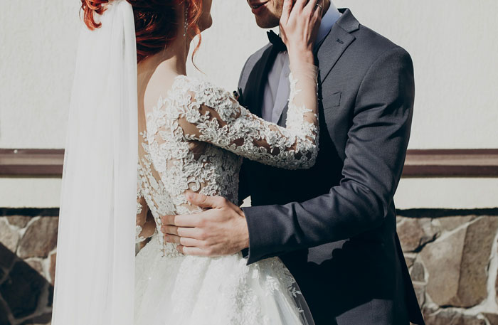 Bride in lace wedding dress and groom in dark suit embrace, representing man gets majorly hurt when wife cancels anniversary trip. Bride in lace wedding dress and groom in dark suit embrace, representing man gets majorly hurt when wife cancels anniversary trip.