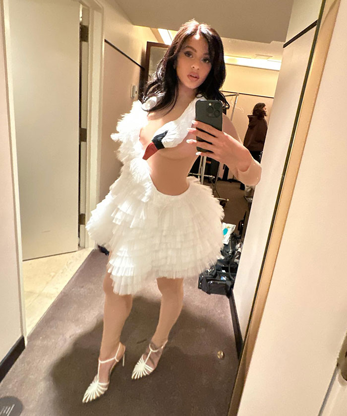 Pop star taking a mirror selfie in a white feather-inspired dress at Paris Fashion Week, showcasing dyed armpit hair.