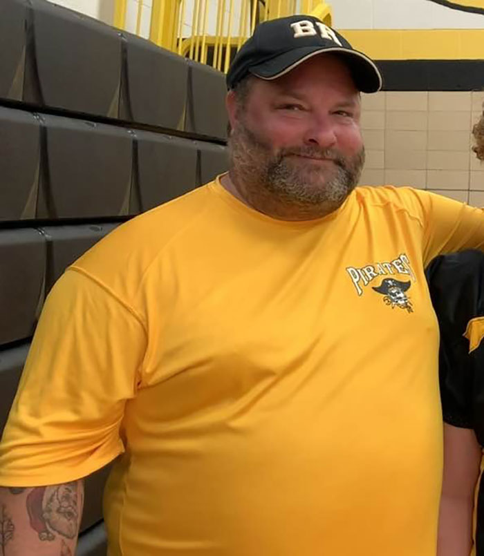 Morbidly obese man wearing a yellow Pirates shirt and black cap, standing indoors near bleachers in a gym.