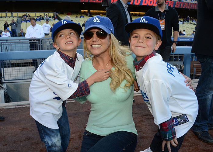 Britney Spears smiling with two children at a baseball stadium, all wearing Dodgers hats and casual outfits.